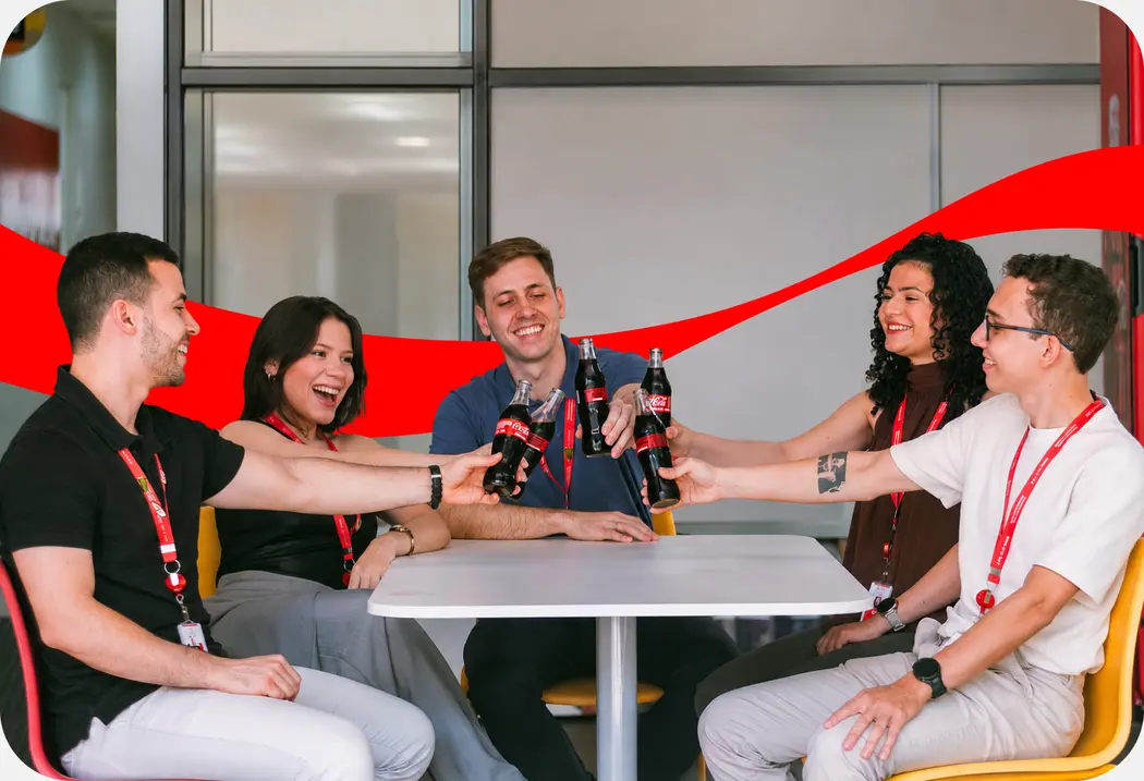 Cinco jovens sentados à mesa de um refeitório brindando com garrafas de Coca-Cola